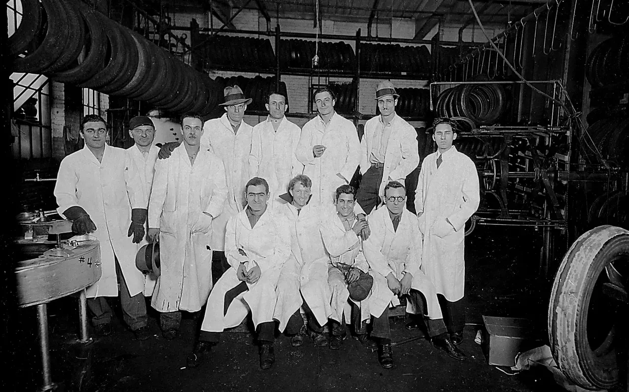 Group of men in white coats posing in a tire workshop with tire racks in the background.