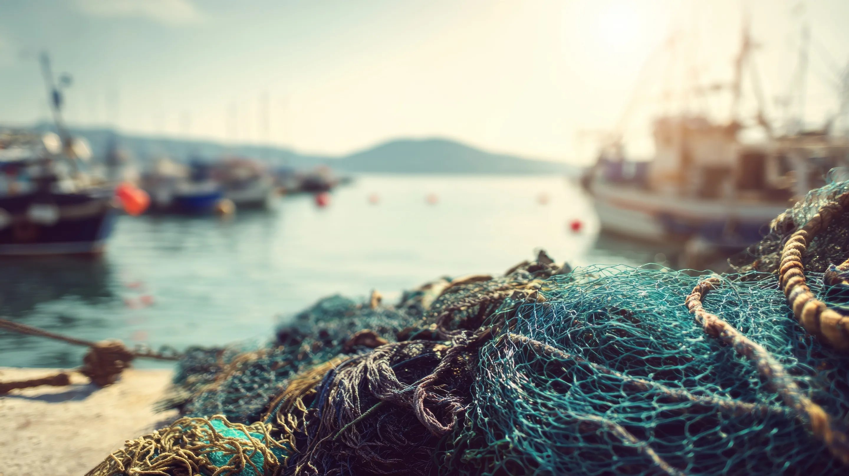 Fishing nets piled on dock with blurred boats and water in the background at a sunlit harbor.