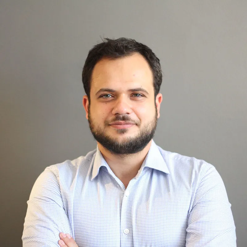 Kolları bağlı, açık mavi kareli gömlek giymiş, hafif gülümseyen sakallı adam.