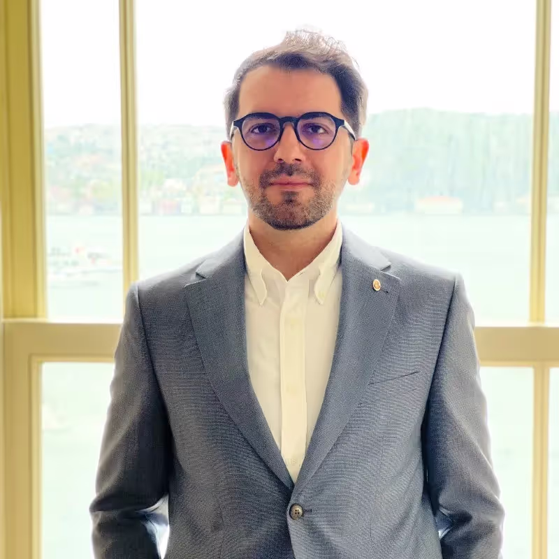 Gözlük takan, takım elbise giyen ve beyaz gömlekli, pencere önünde poz veren yetişkin erkek.