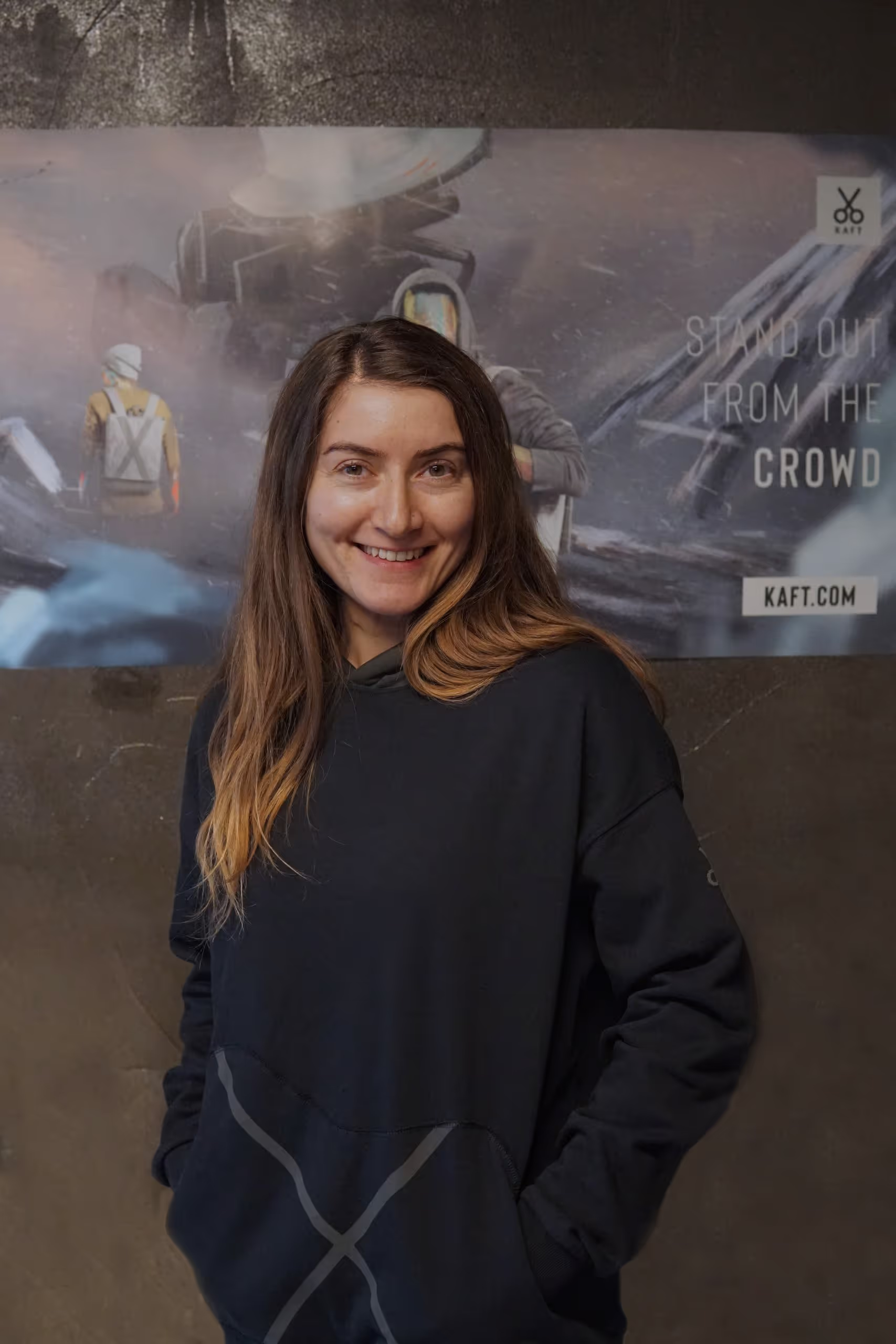 Gülümseyen uzun kahverengi saçlı kadın, koyu renk kapüşonlu sweatshirt giymiş ve elleri ceplerinde duruyor.