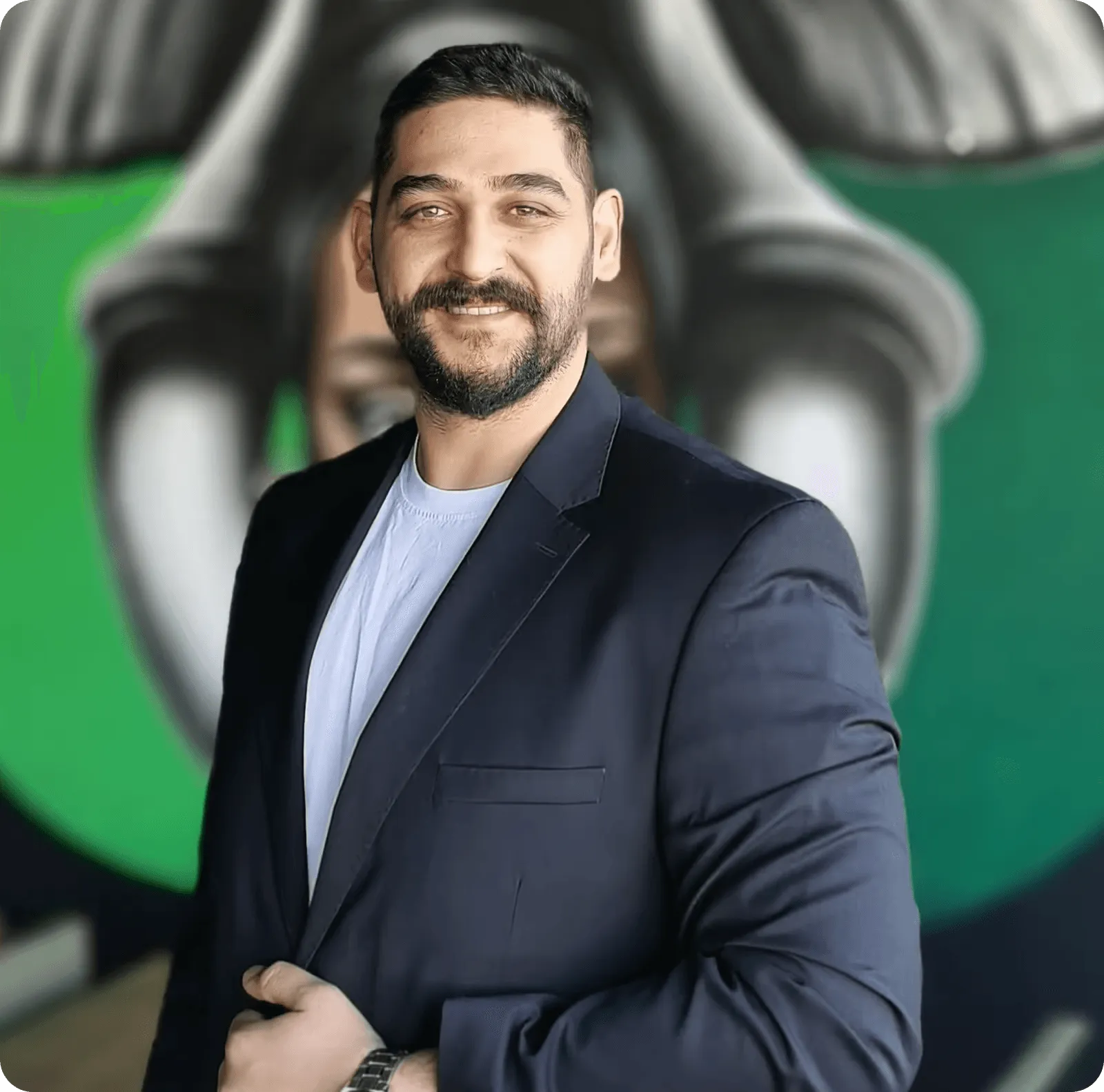 Gülümseyen, beyaz tişört ve koyu blazer ceket giymiş, arkada yeşil ve gri graffiti bulunan adam.