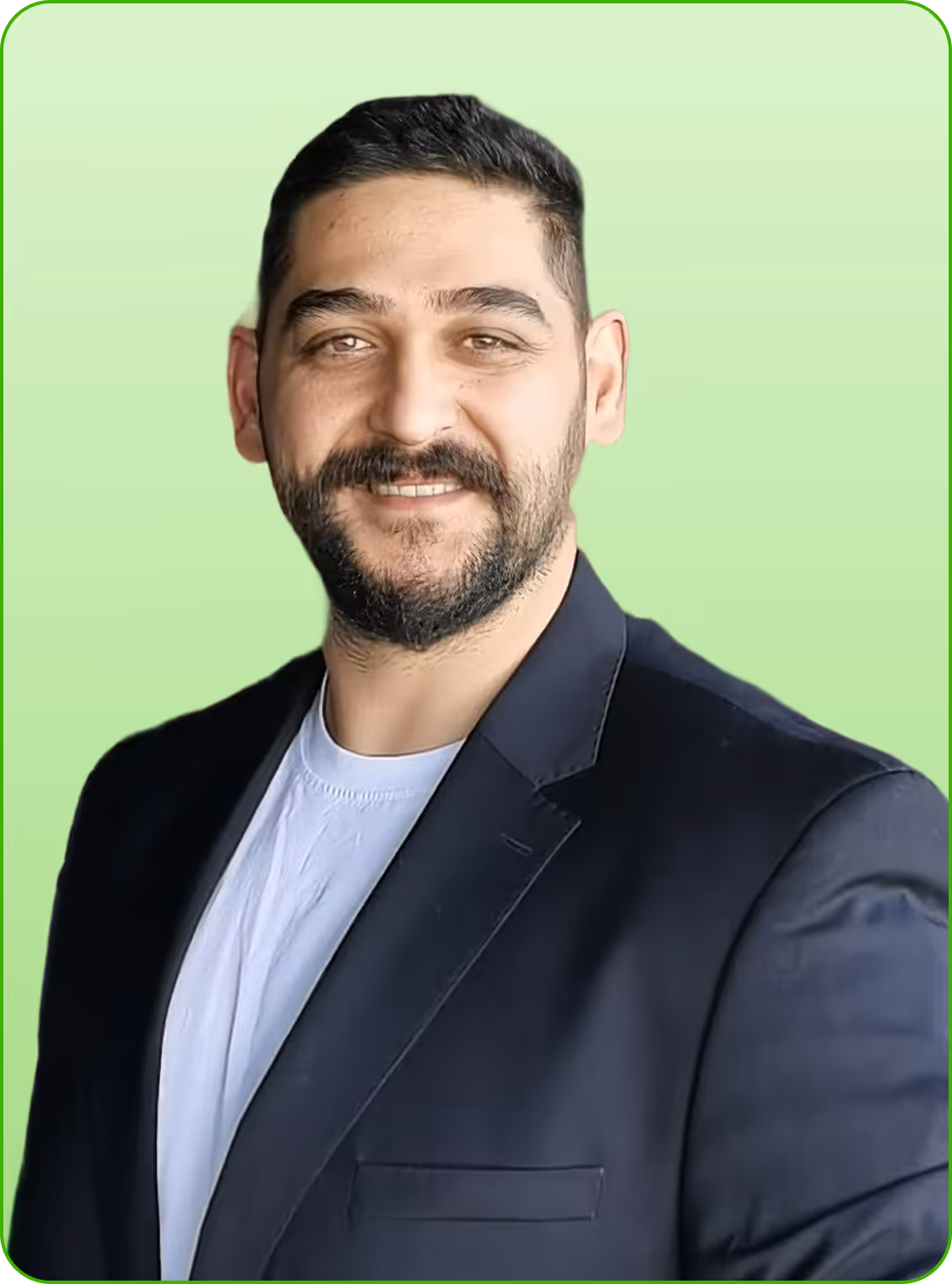 Kısa sakallı, koyu saçlı ve koyu ceketli, açık renk tişört giymiş gülen adam.