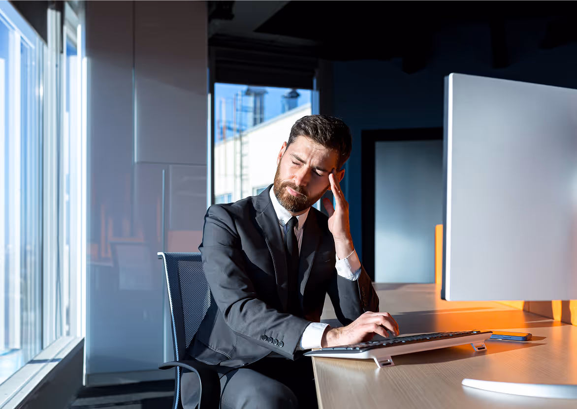 Man in pak zit aan bureau met hand aan hoofd, lijdt aan hoofdpijn in modern kantoor.