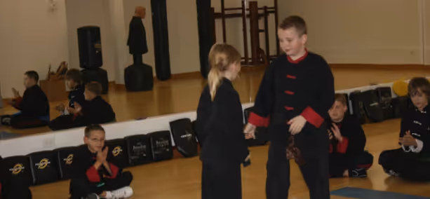 Children in black martial arts uniforms practicing in a gym with padded equipment along the wall.