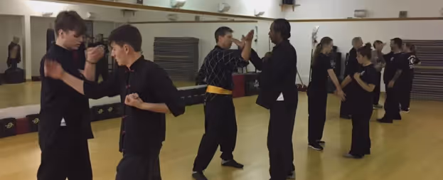 Several pairs practicing martial arts techniques inside a gym with wooden floor and mirrors.