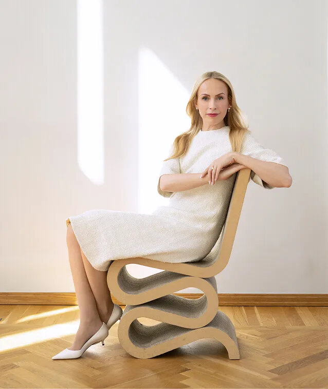 Blonde woman in a white dress sitting on a modern wavy chair in a sunlit room with wooden floor.