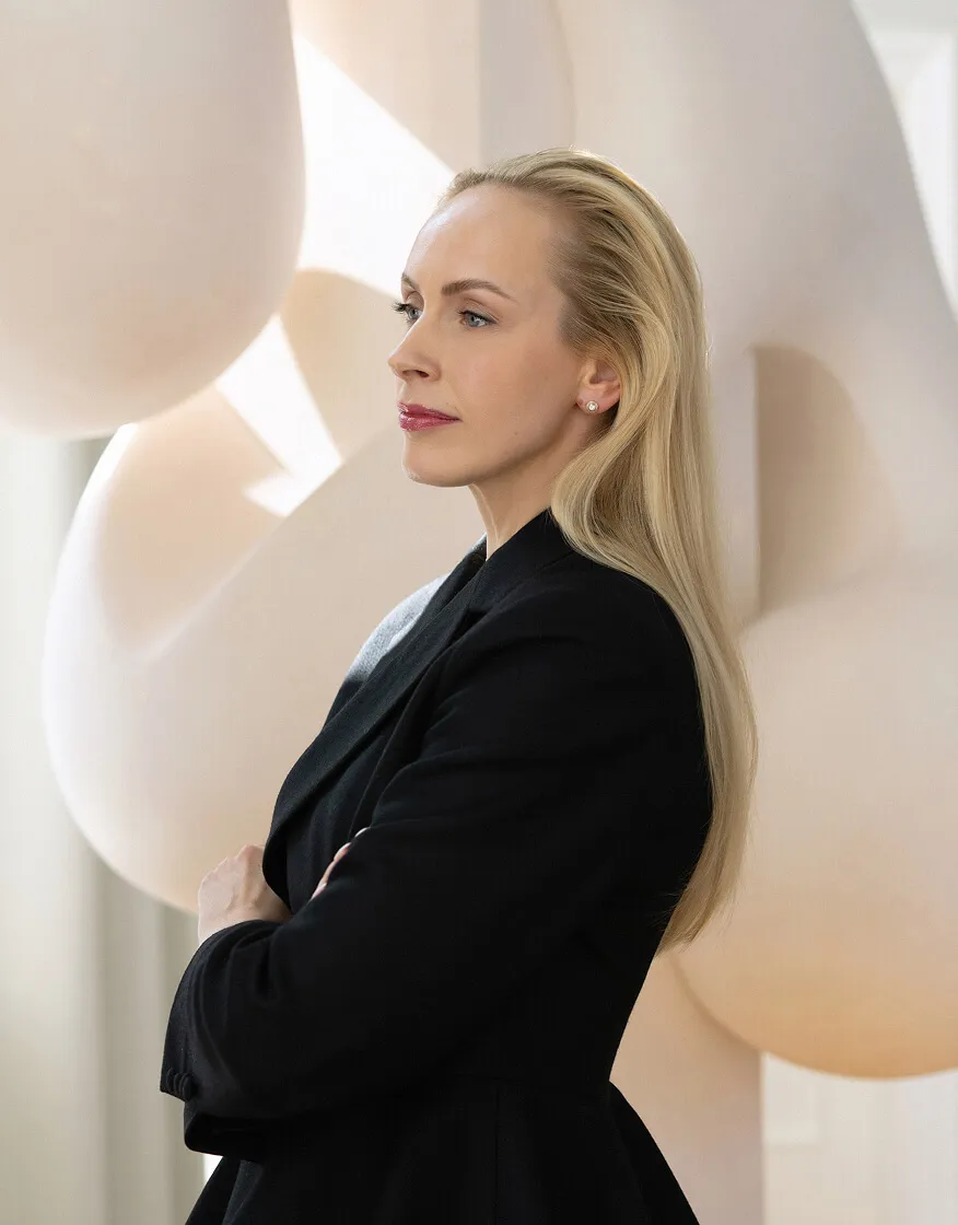 Confident blonde woman in black jacket with arms crossed, looking to the side against a sculptural background.
