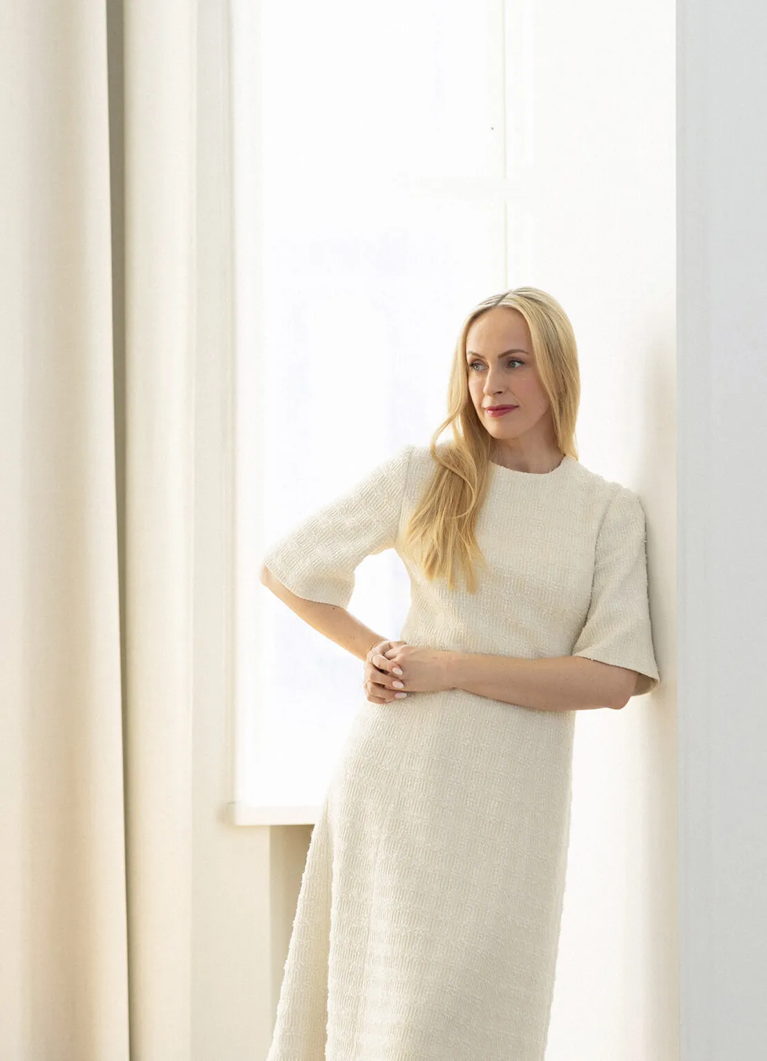 Blonde woman in a textured cream dress leaning against a white wall by a window with curtains.