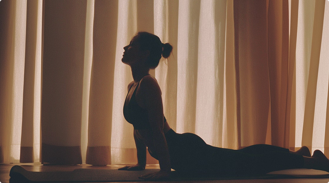 Silhouette of a woman doing a yoga cobra pose on a mat in front of light curtains.