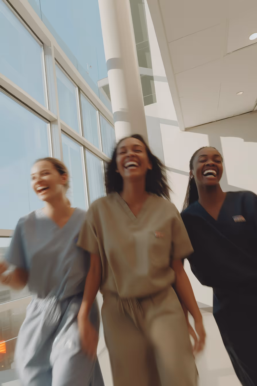 Three female healthcare workers in scrubs laughing and walking in a bright hallway with large windows.