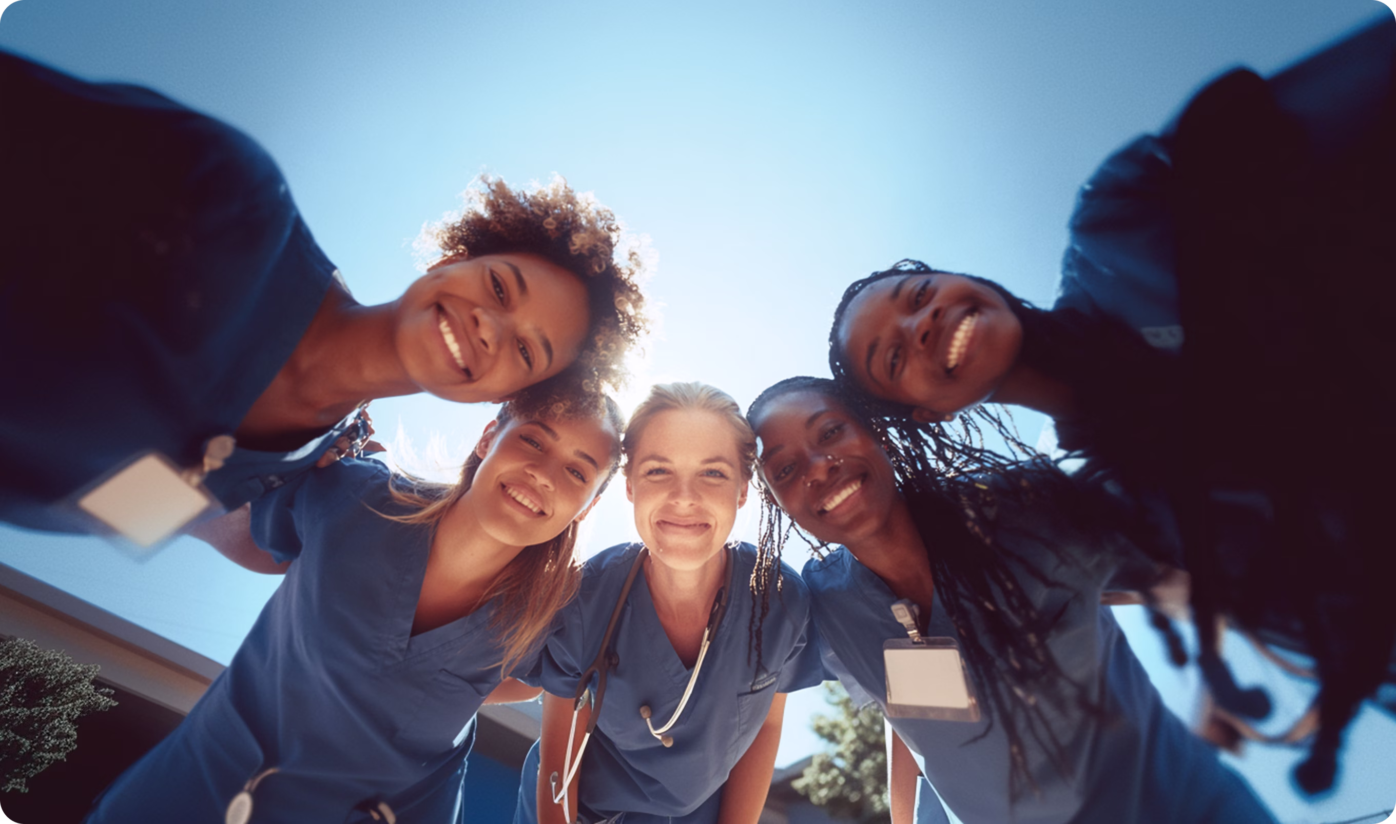 Group of five smiling medical professionals in blue scrubs looking down at the camera outdoors.