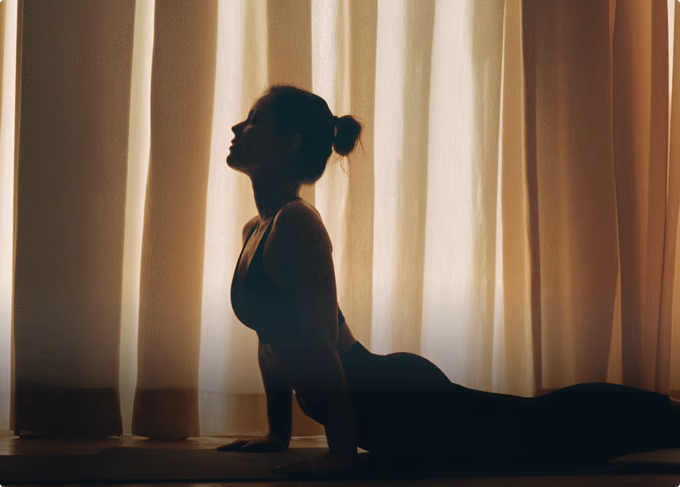 Silhouette of a woman doing yoga in upward-facing dog pose against a curtain-lit background.