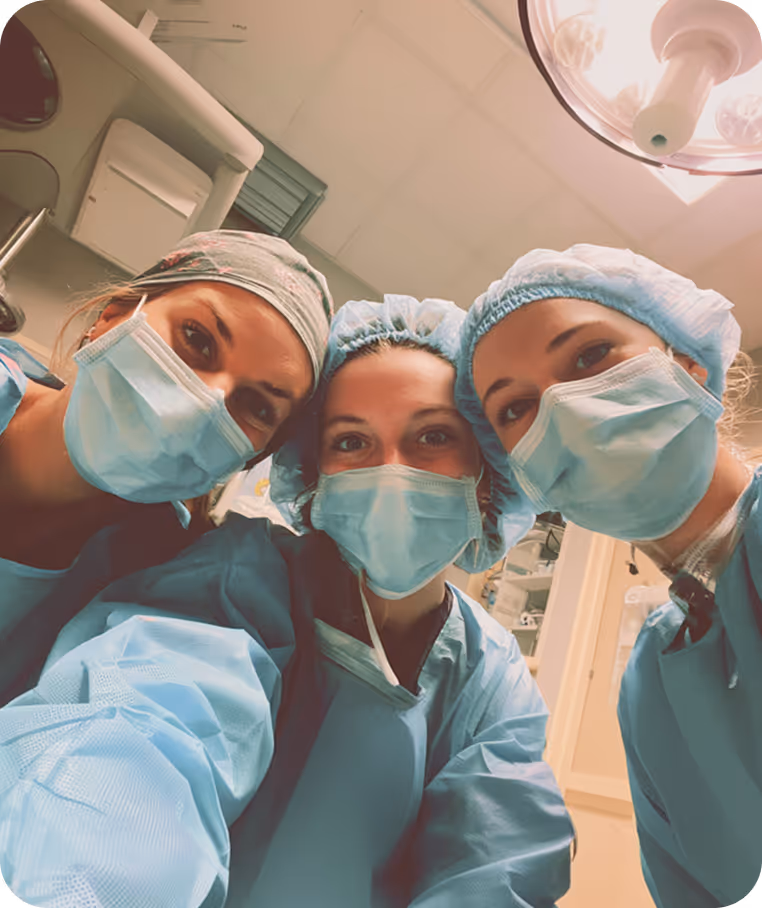 Three healthcare workers in scrubs, masks, and caps looking at the camera in a medical setting.