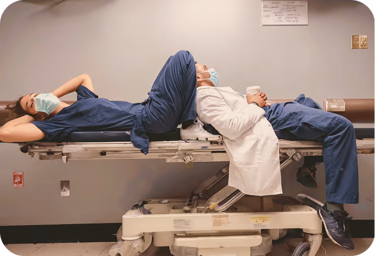 Two healthcare workers resting back-to-back on a hospital stretcher wearing scrubs and masks.