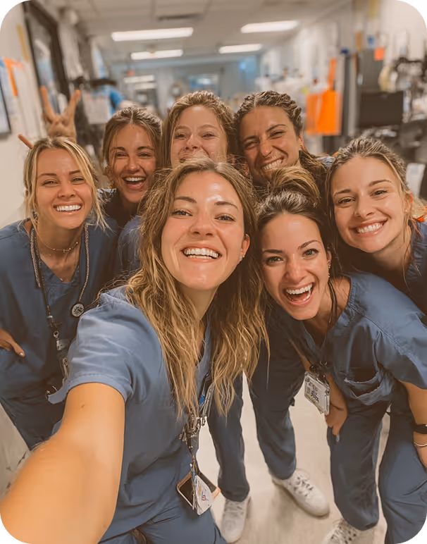 Seven smiling healthcare workers in blue scrubs taking a group selfie in a medical hallway.