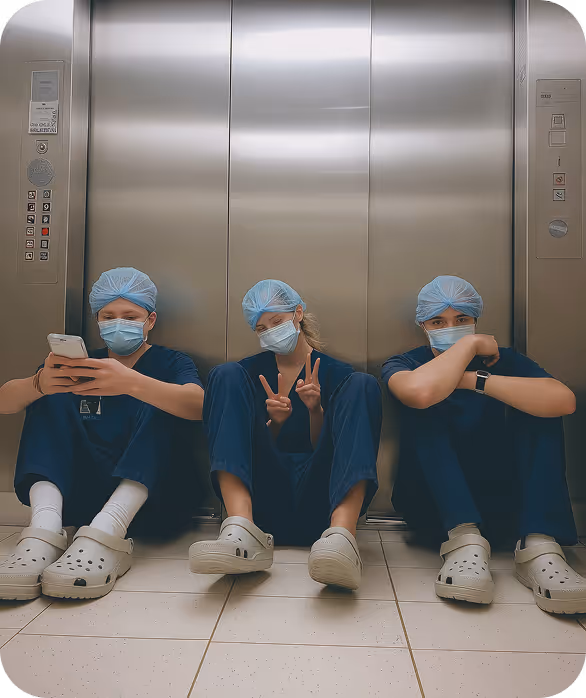 Three healthcare workers sitting on the floor in front of an elevator, wearing scrubs, masks, and surgical caps.