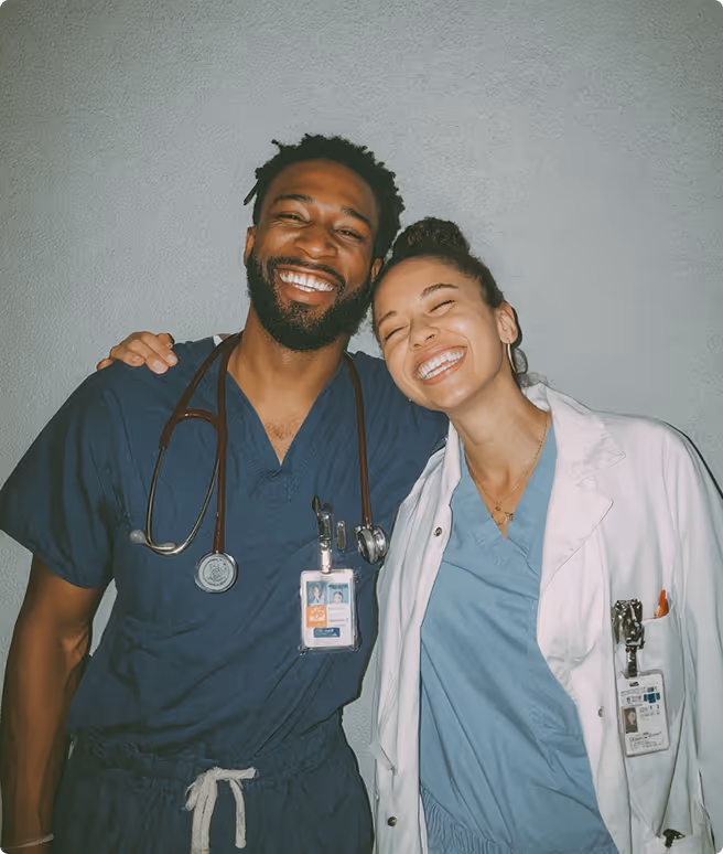 Two smiling healthcare workers in scrubs and a white coat with stethoscopes and IDs.