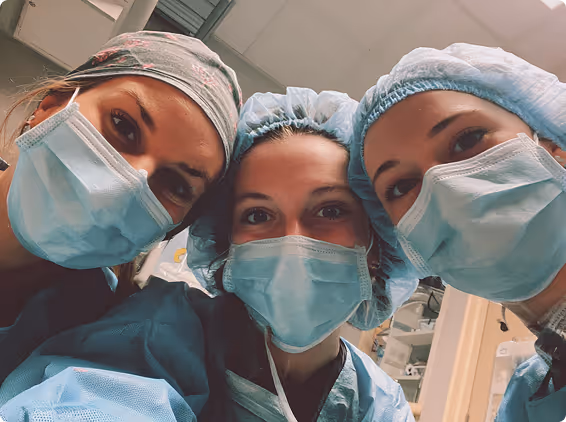 Three healthcare workers wearing surgical masks, caps, and scrubs posing for a selfie indoors.