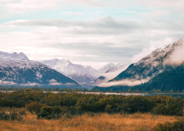 Snow-capped mountains with clouds and fog over a forested valley and golden grassland.