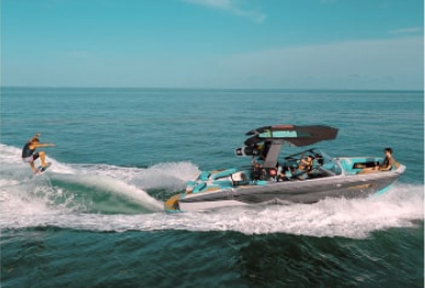 Wakeboarder riding on a wave behind a speedboat on a calm, clear blue sea.