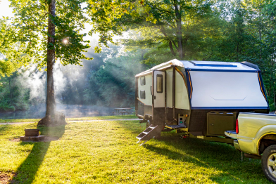 Sunlight filtering through trees onto a parked RV camper and grassy campground with fire pit.