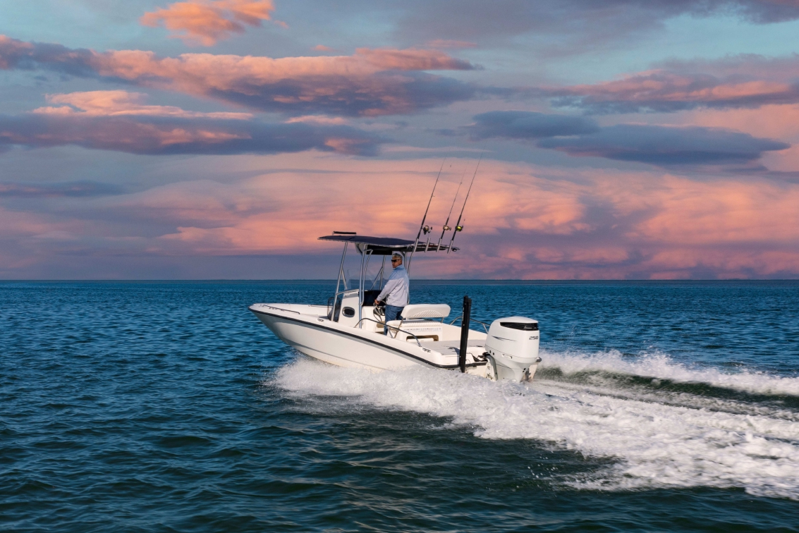 Person steering a white fishing boat with rods on calm water under a pink and blue sky.