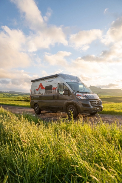 Camper van parked on a dirt road in a green field under a partly cloudy sky at sunset.