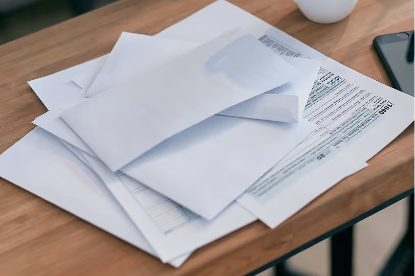 Stack of white envelopes and tax form 1040 on a wooden table with a smartphone and a cup nearby.