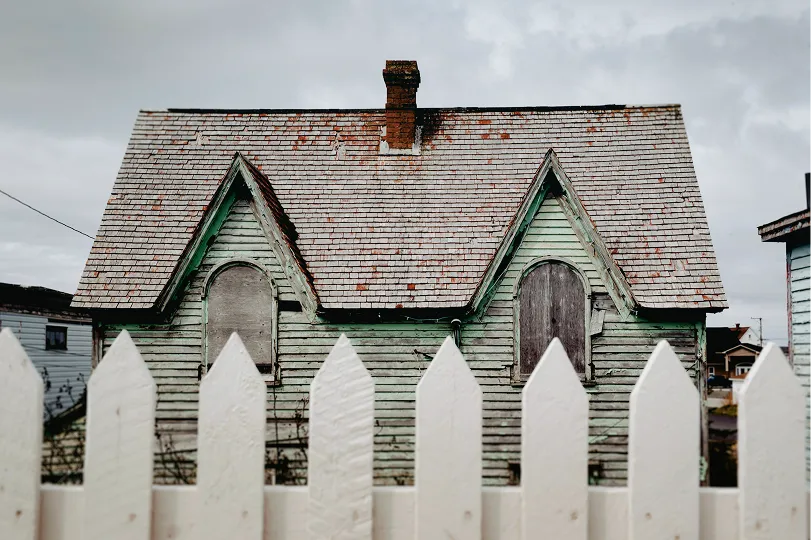Old, weathered green wooden house with boarded arched windows behind a white picket fence under an overcast sky.