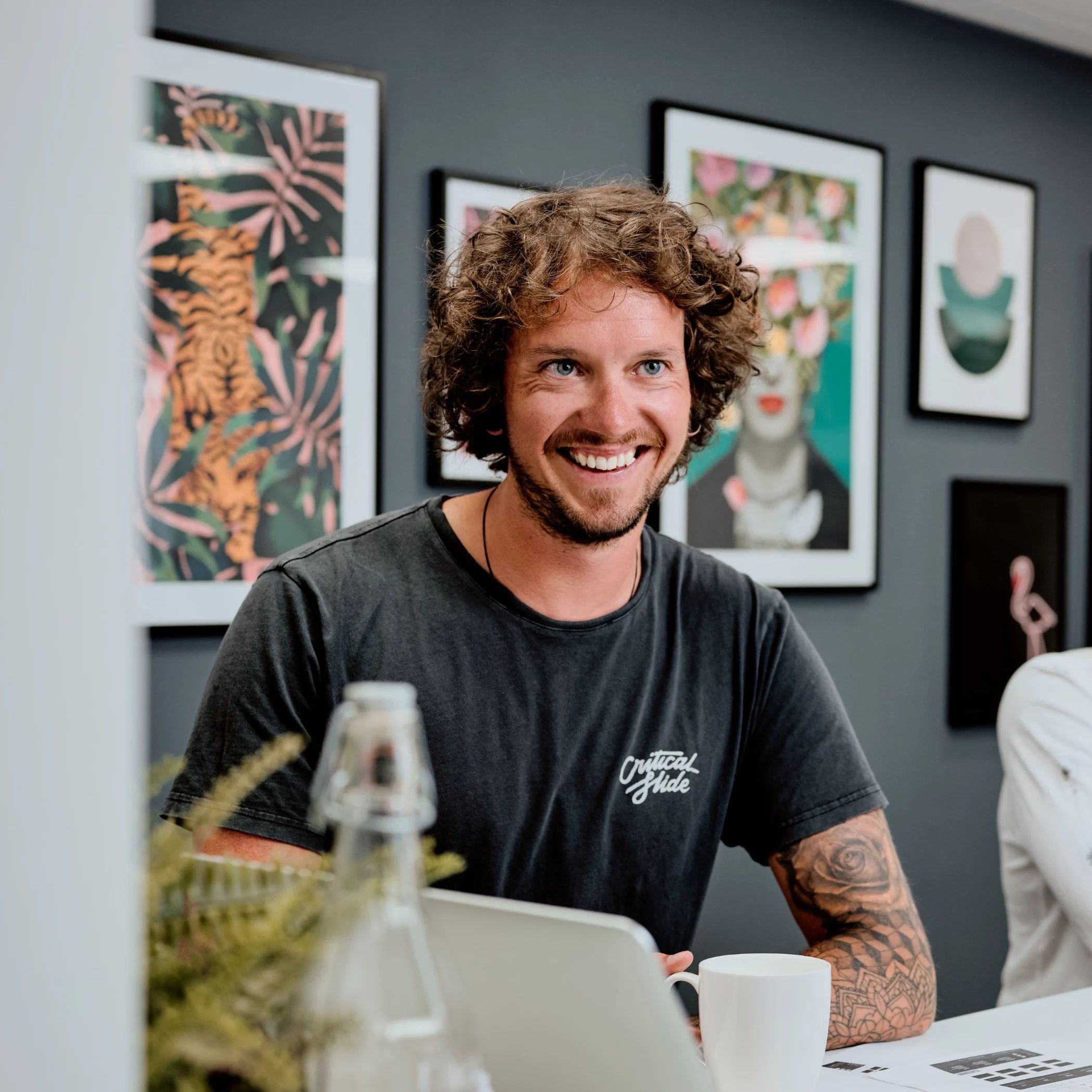 Smiling man with curly hair and tattooed arm sitting at a table with a laptop, coffee cup, and artistic framed pictures on a gray wall behind him.