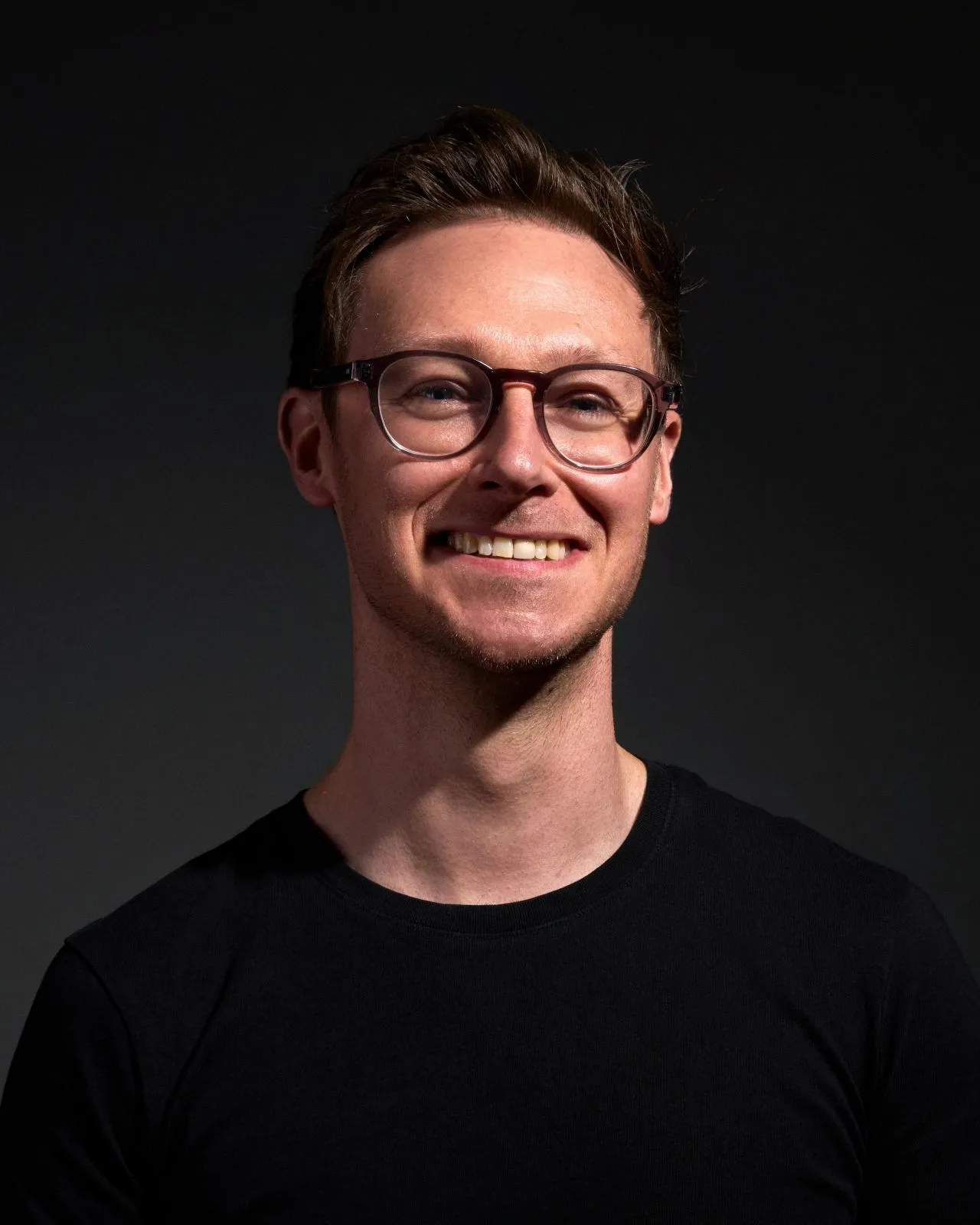 Smiling man with short brown hair, glasses, and black shirt against a dark background.