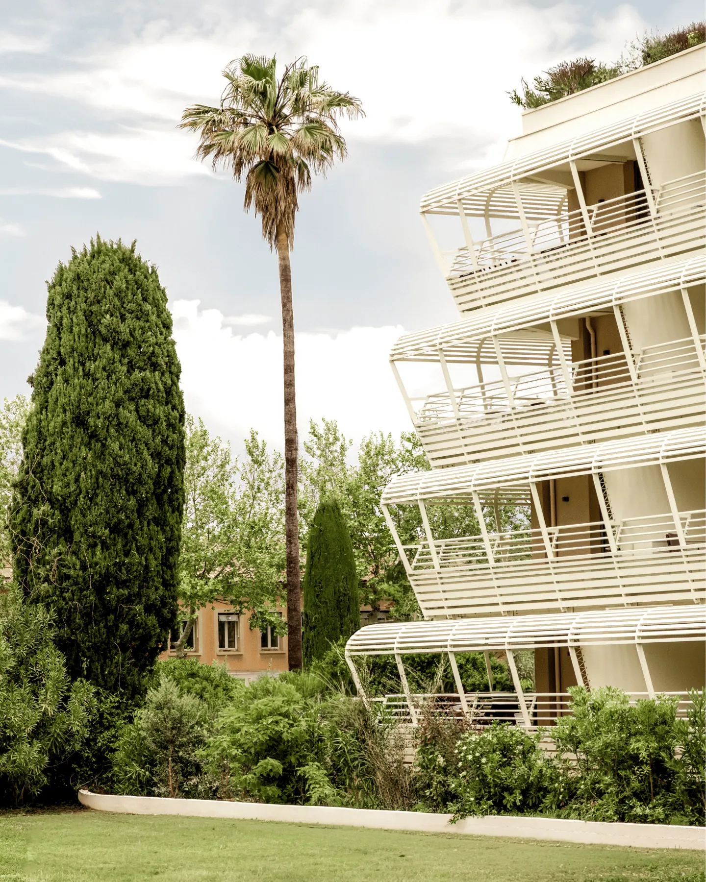Modern building with arched white balconies and palm trees surrounded by greenery under a cloudy sky.