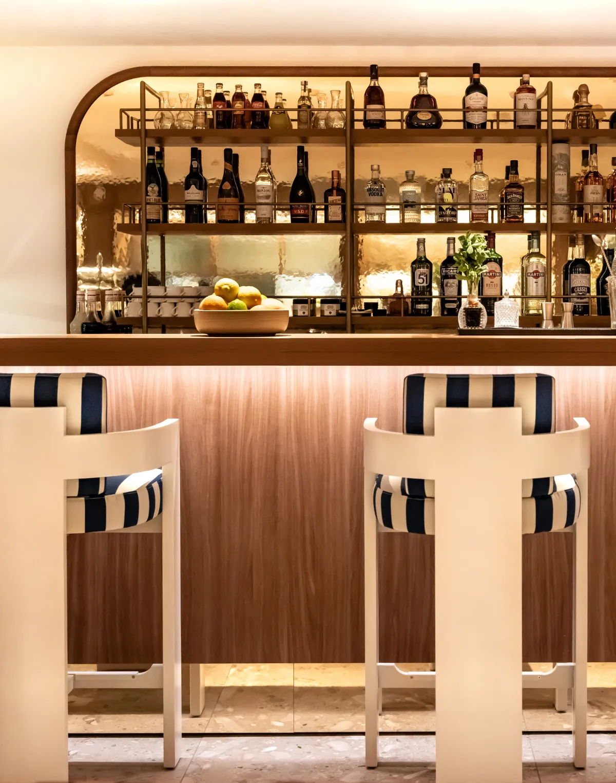 Bar with white stools with blue and white striped cushions, bottles and fruit on the counter.