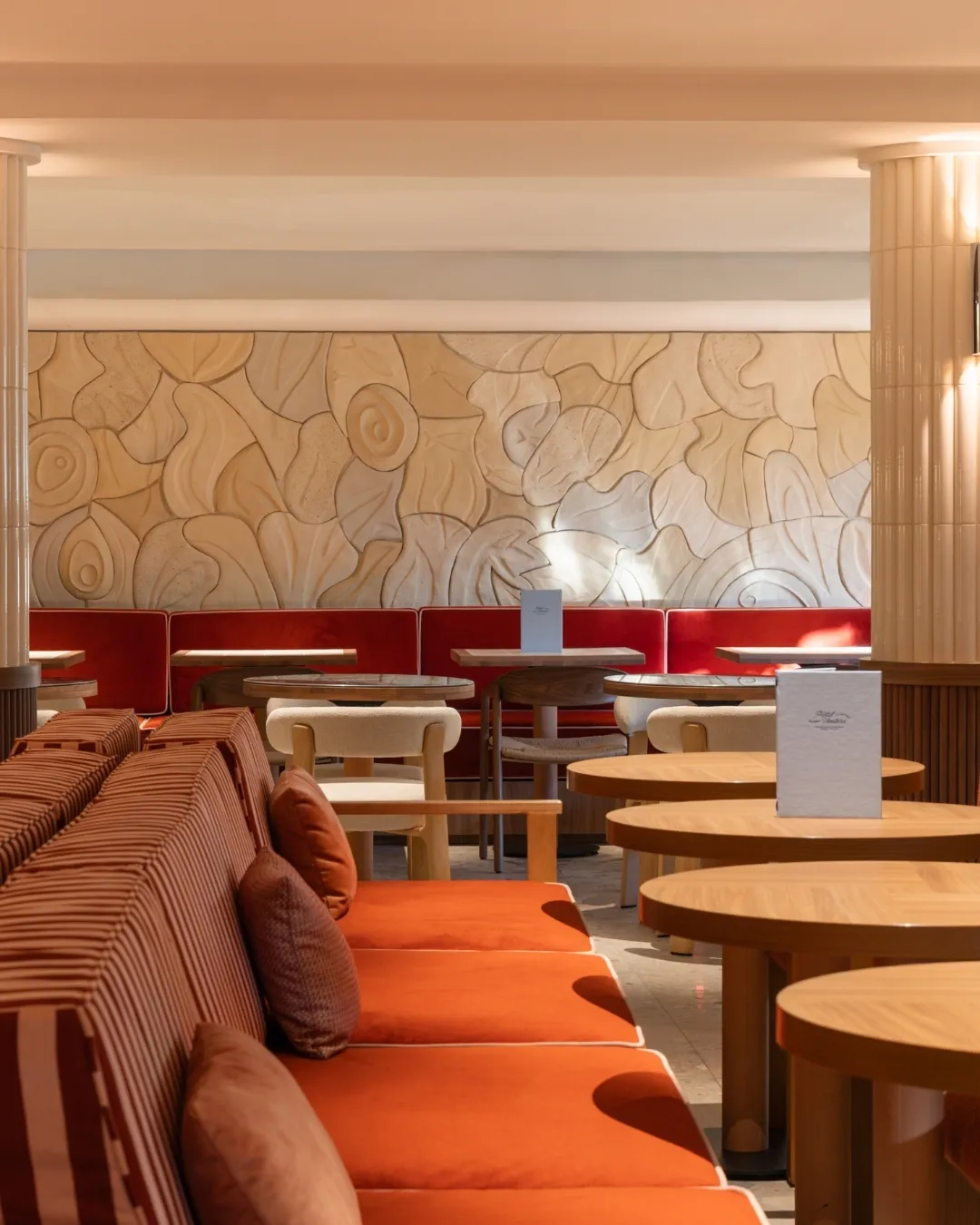 Restaurant avec canapés orange, tables en bois rondes et mur décoré de formes organiques beige.