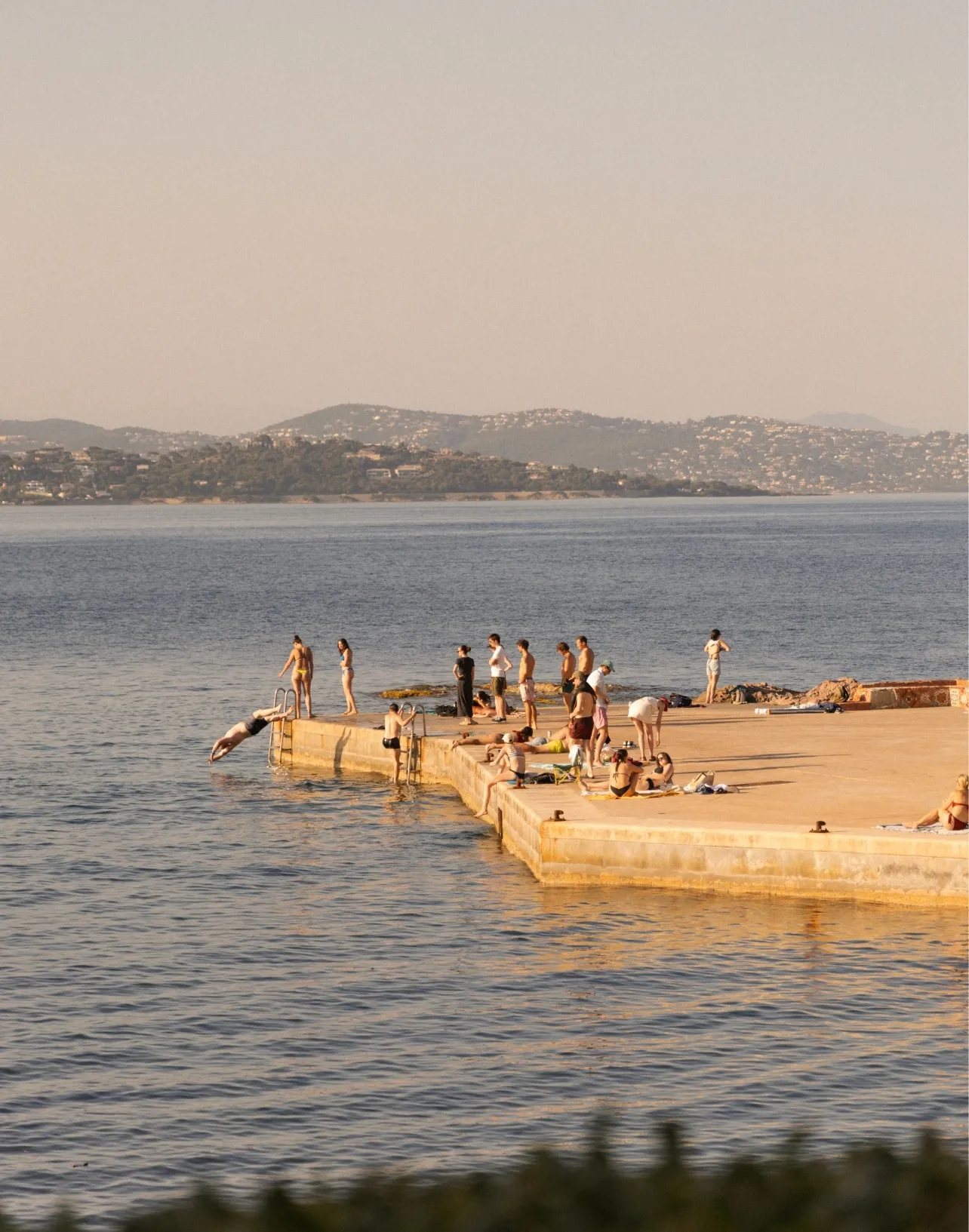 Des gens se baignent et bronzent sur une jetée au bord de la mer avec des collines en arrière-plan.
