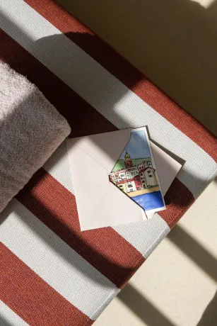 Envelope with a Mediterranean village illustration resting on a terracotta and white striped deckchair in the sun