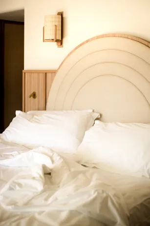 Unmade bed with white sheets and pillows in front of a light wood headboard with Art Deco curves and a rattan wall sconce