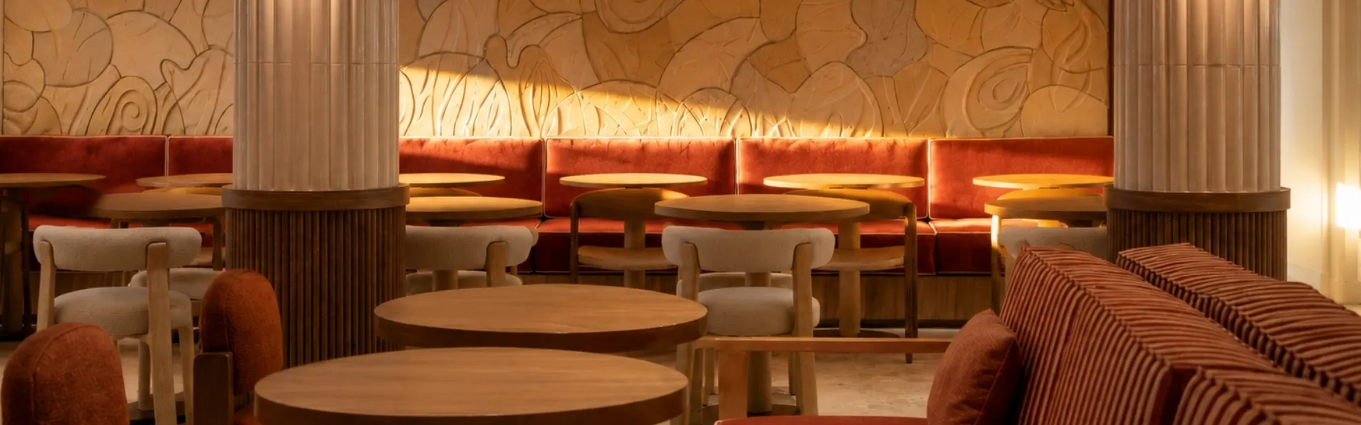 Elegant dining room in warm tones with terracotta velvet banquettes, round wooden tables, and white columns in front of a backlit sculpted wall