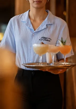 Waiter in a blue shirt carrying a tray of colorful cocktails in glasses, including one peach drink and two creamy white drinks