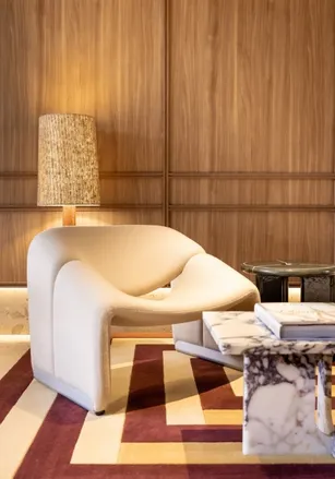 Designer living room with a white, rounded armchair, a marble coffee table, and a rattan lamp, against a backdrop of wood paneling, and a burgundy and beige geometric rug.
