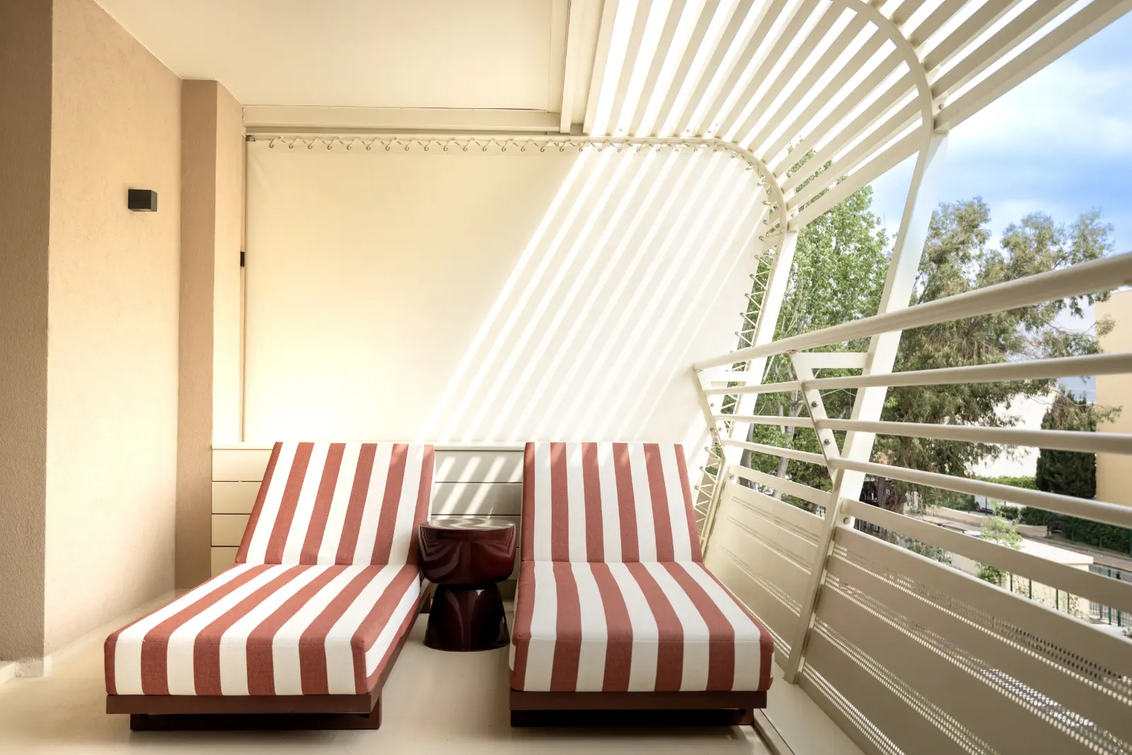 A sunny hotel balcony with two terracotta-and-white striped sun loungers under a pergola with white slats