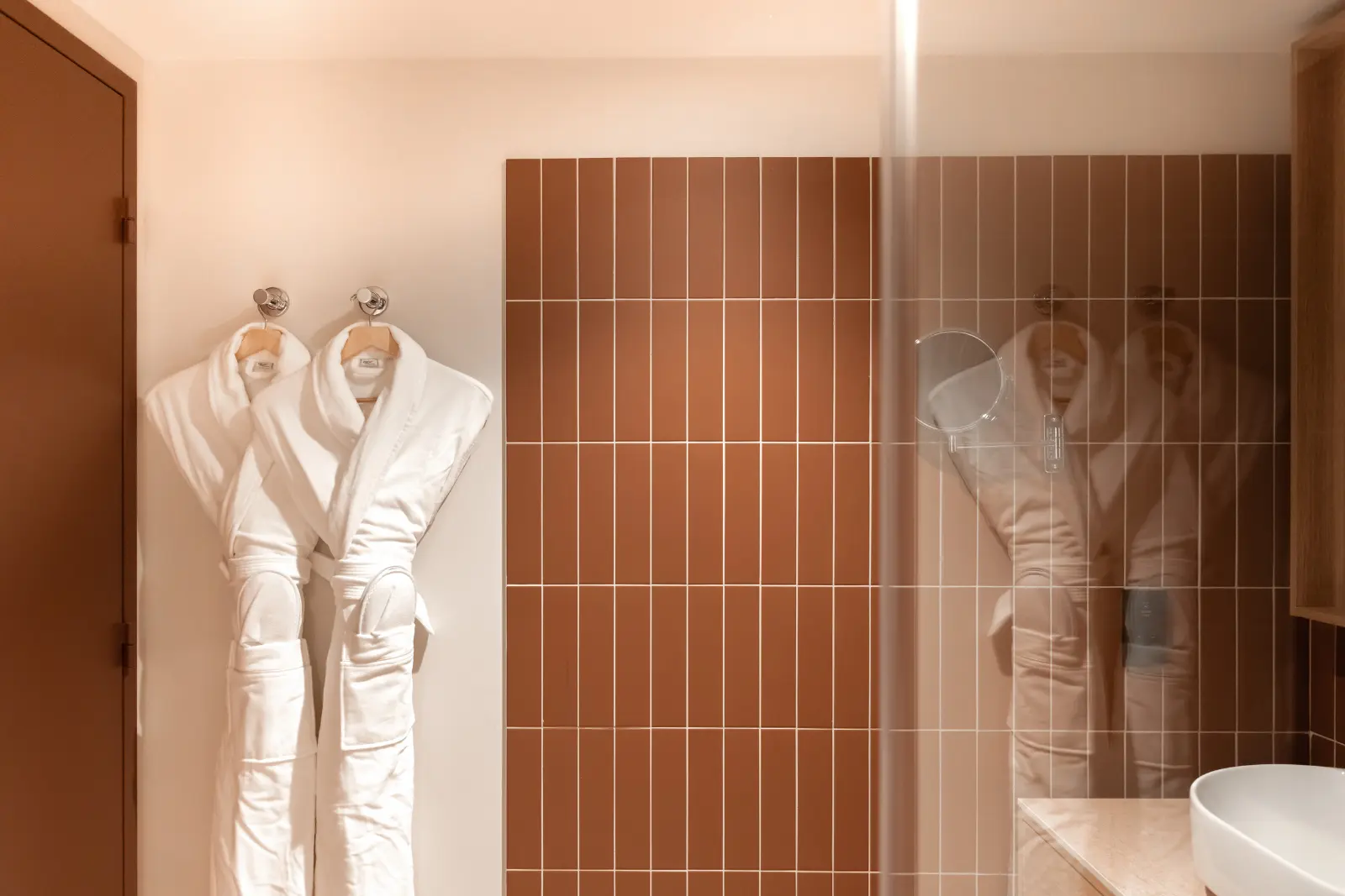 A hotel bathroom with two white bathrobes hanging on a wall of warm-toned brown tiles