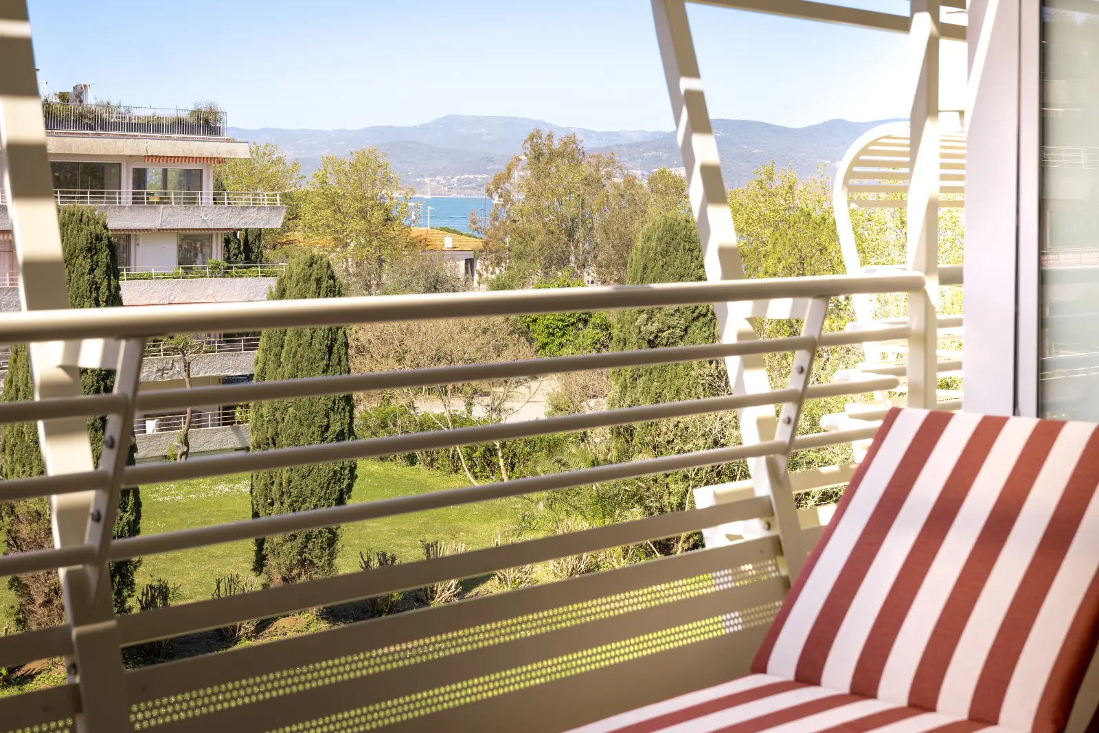 A hotel balcony with a red-and-white striped chair, a pergola with white slats and a view of the hills