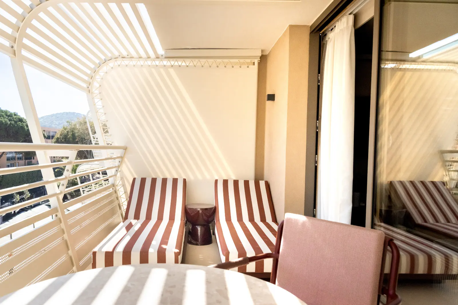 A hotel balcony with two terracotta-and-white striped sun loungers beneath a pergola with curved white slats