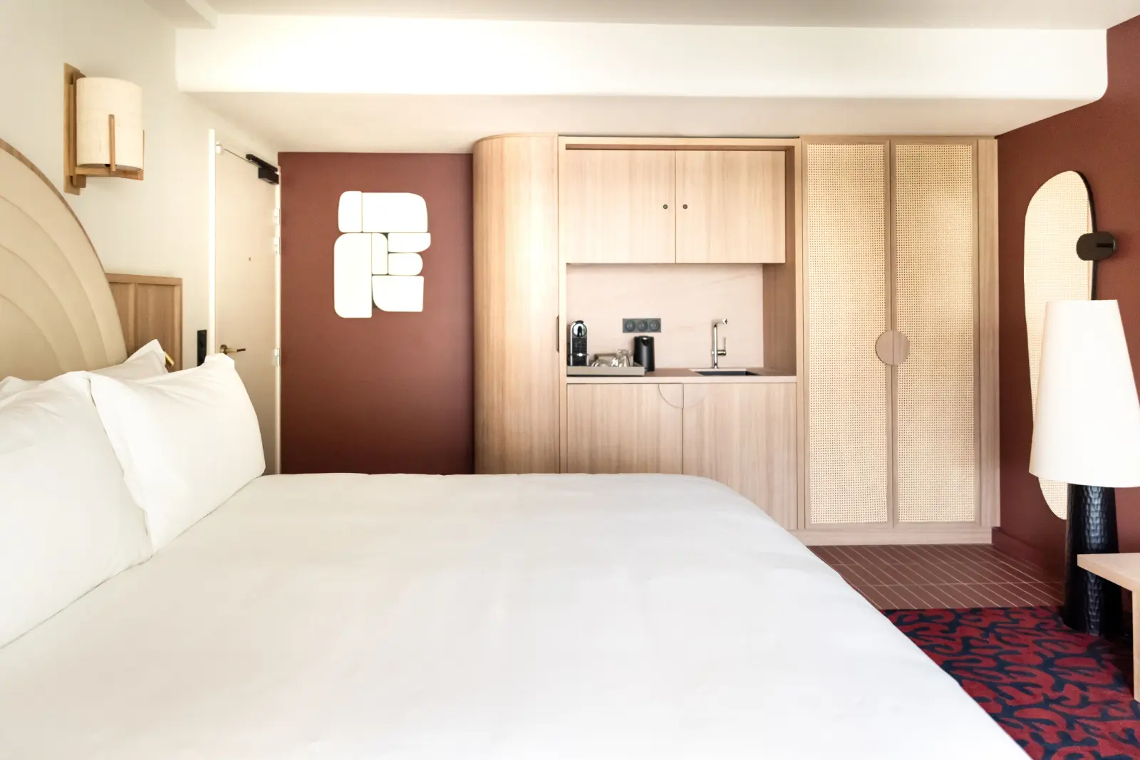 Hotel room with a double bed, a light-coloured rounded headboard, a red rug with organic patterns, and a burgundy door