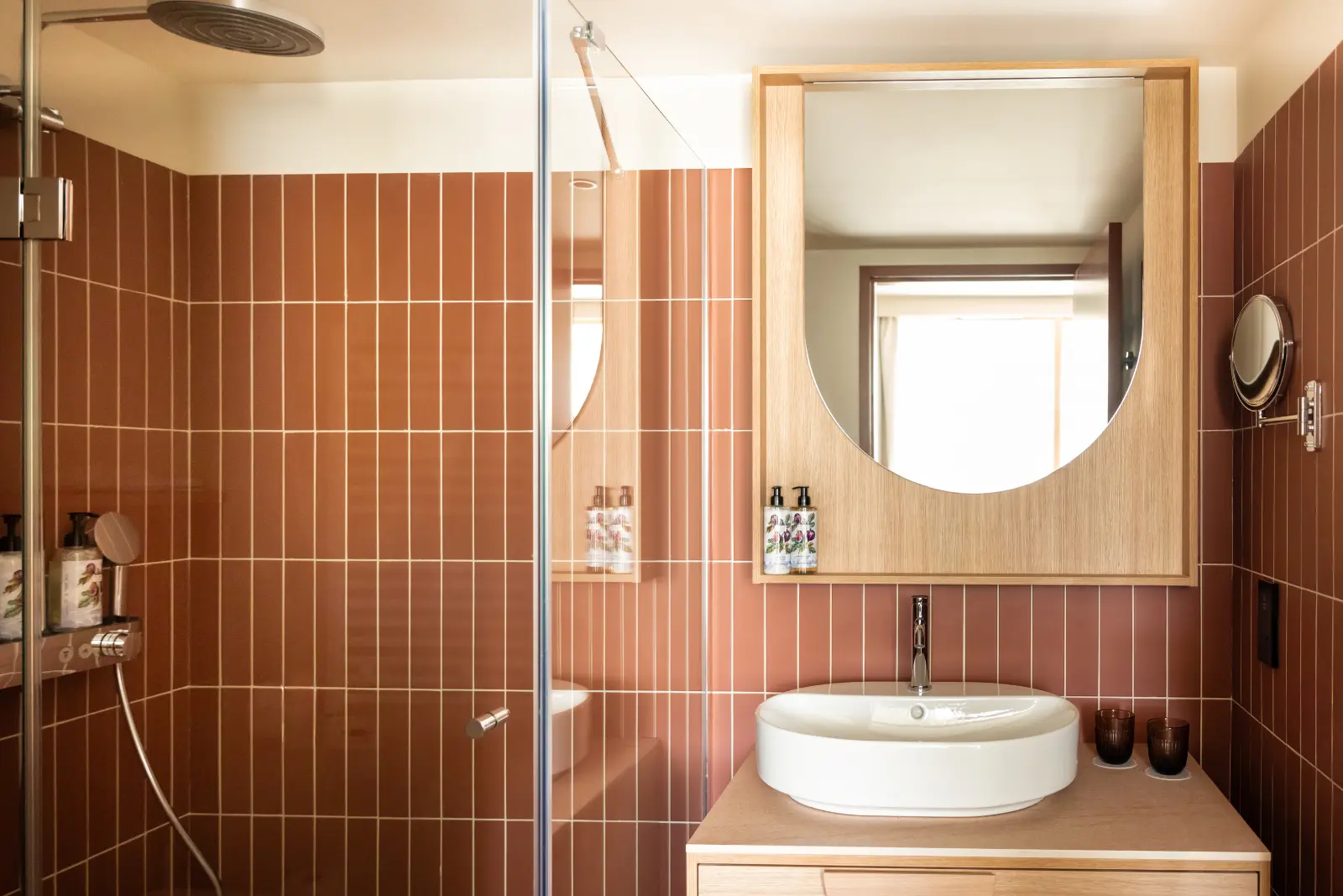 Hotel bathroom with a walk-in shower, brown tiles, a round white washbasin and an oval mirror
