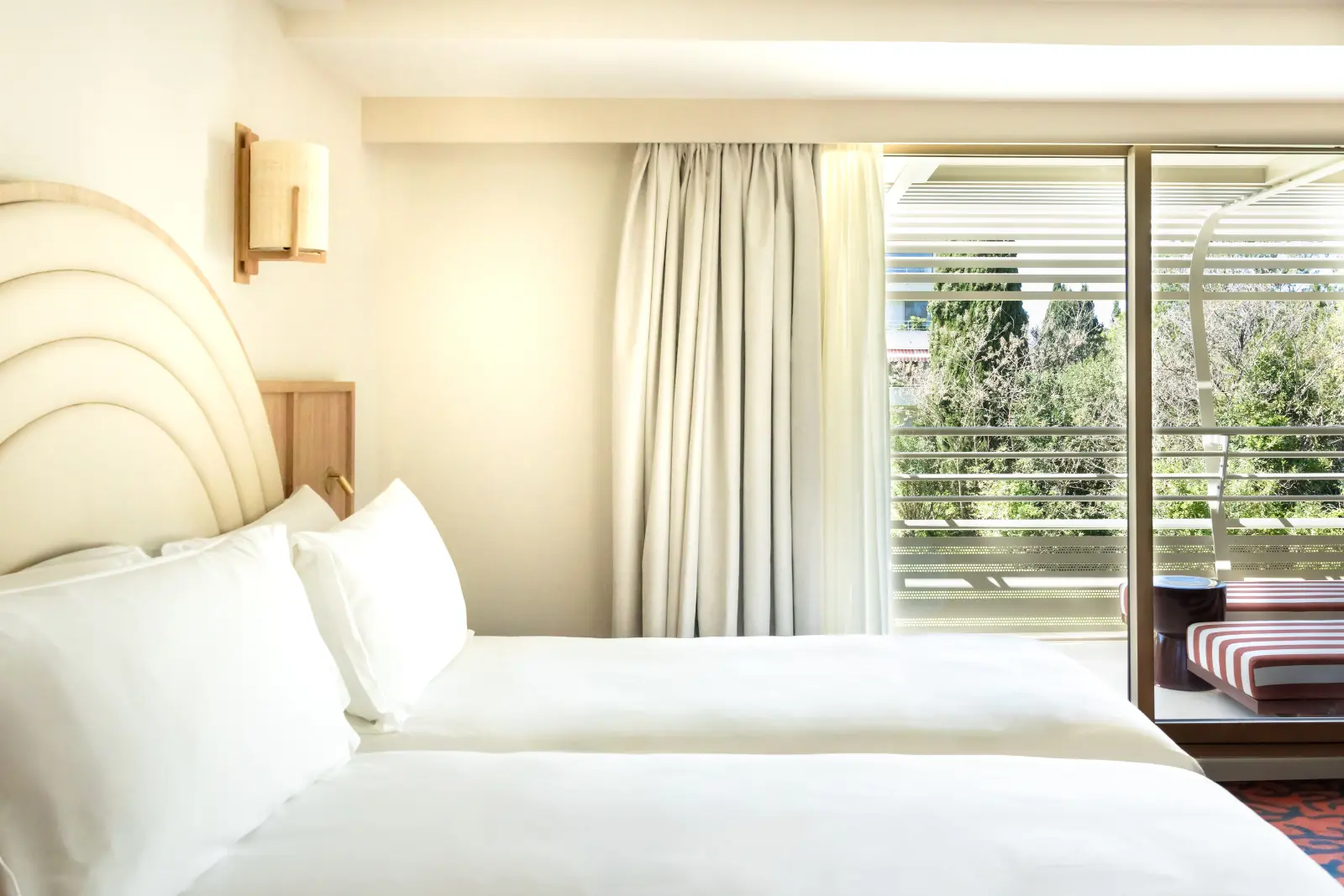 A bright hotel room with a double bed, a white curved headboard and a floor-to-ceiling window overlooking the greenery