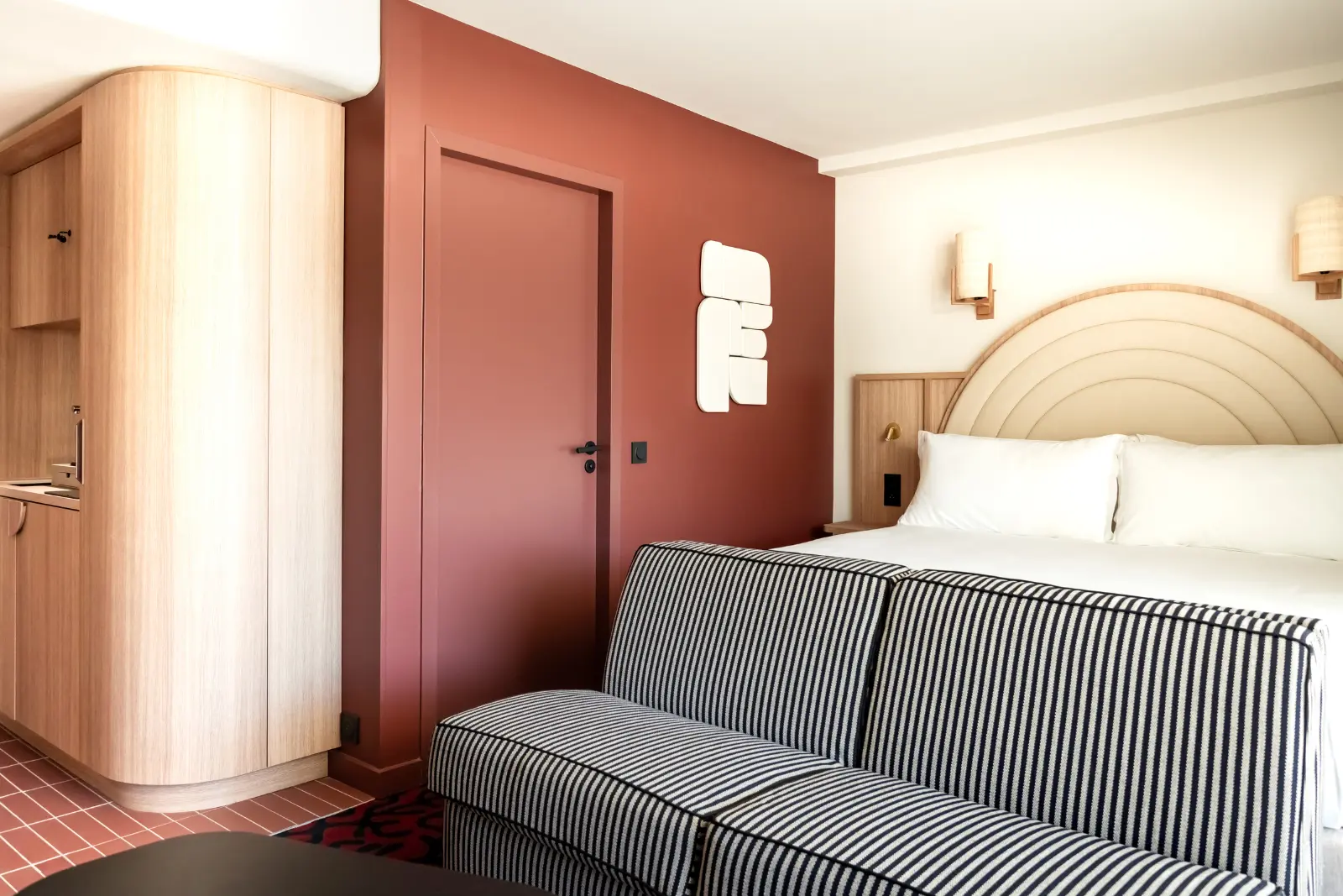 Hotel room with a double bed, a wooden headboard featuring concentric arches, terracotta walls and a black-and-white striped sofa