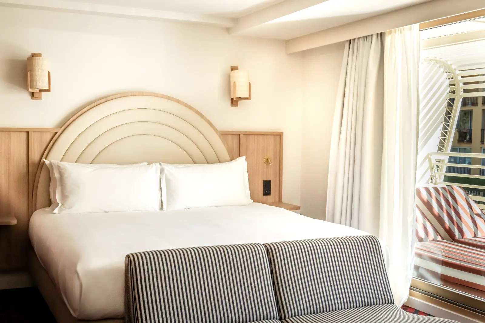 Hotel room with a double bed dressed in black-and-white striped bed linen, a wooden headboard featuring concentric arches, and rattan wall lights